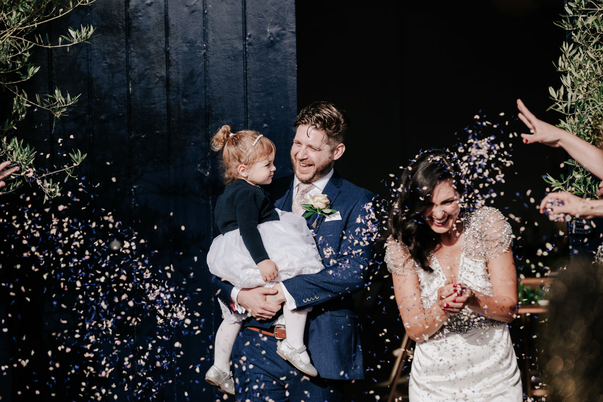 groom holding his daughter and bride leaving chainstore into confetti