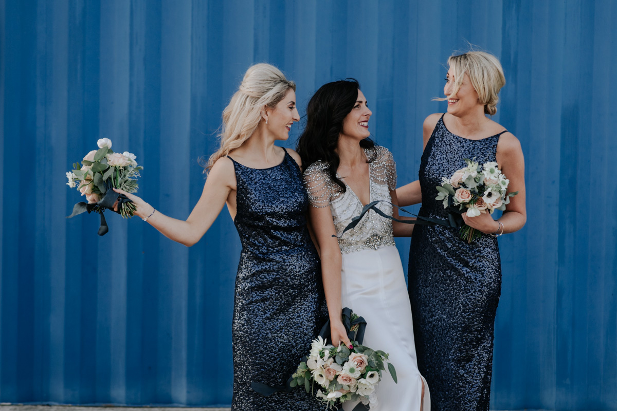 bride with bridesmaids in blue sequin dresses against blue sea containers