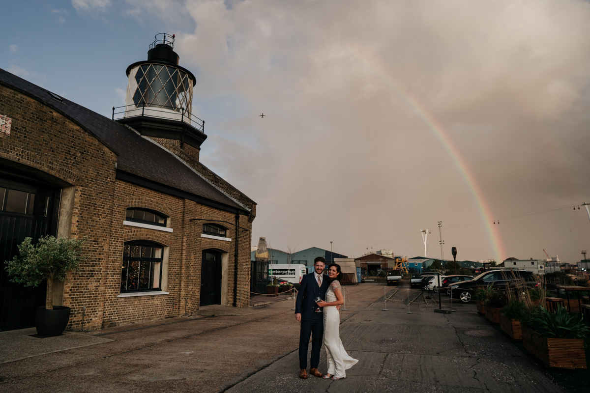 bride and groom with a rainbow in sky and trinity buoy wharf wedding venue and lighthouse