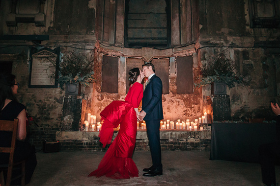 bride in red designer wedding dress kisses groom at candle lit winter asylum chapel wedding. photo by london wedding photographer Joasis
