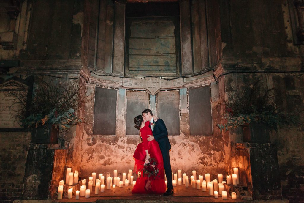 groom kissing bride in designer red wedding dress on altar of asylum chapel surrounded by candles