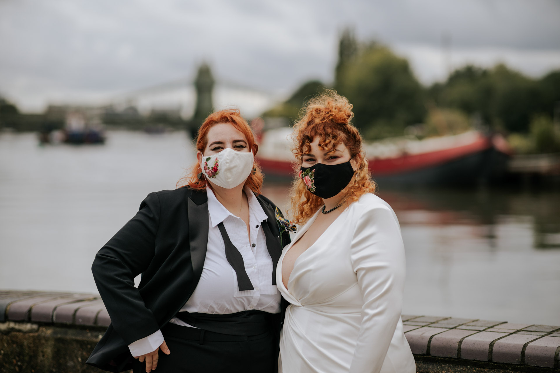 two brides in masks posing for a riverside portrait
