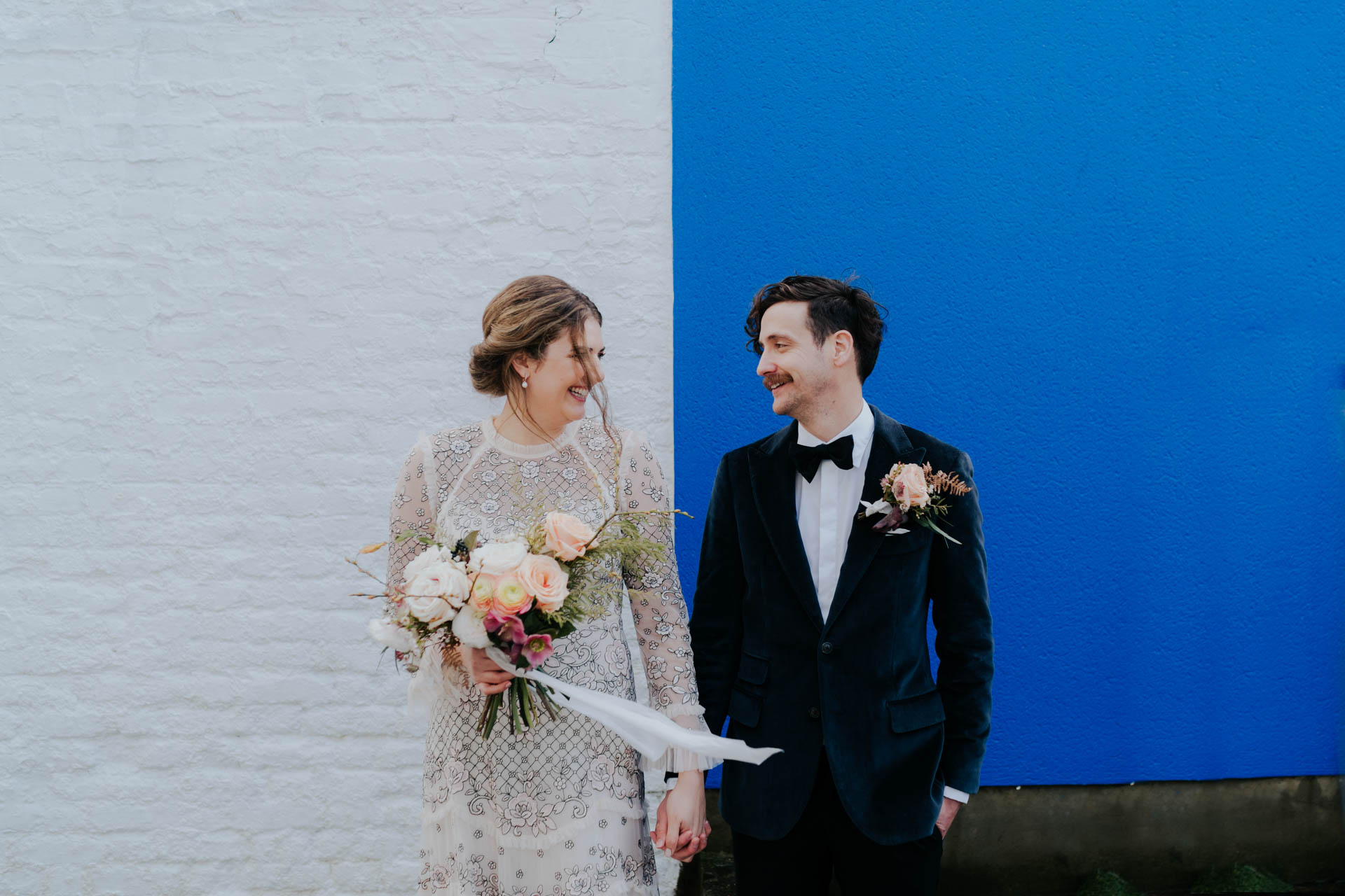 bride and groom looking at each other and smiling against white and blue wall