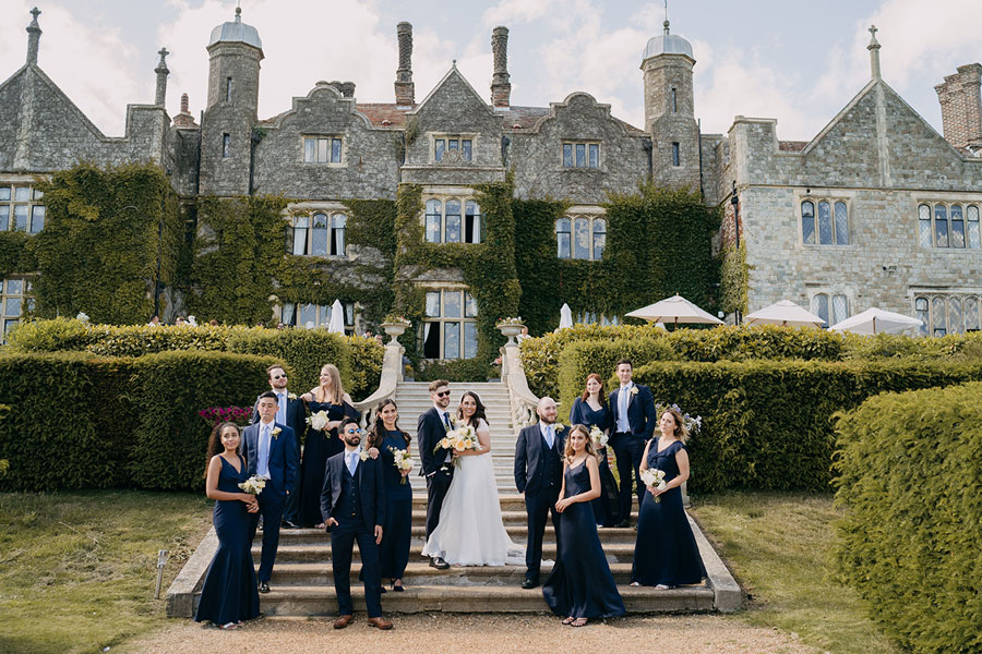 Wedding party posed outside of Eastwell Manor. Bridesmaids in navy long dresses and groomsmen in navy suits.