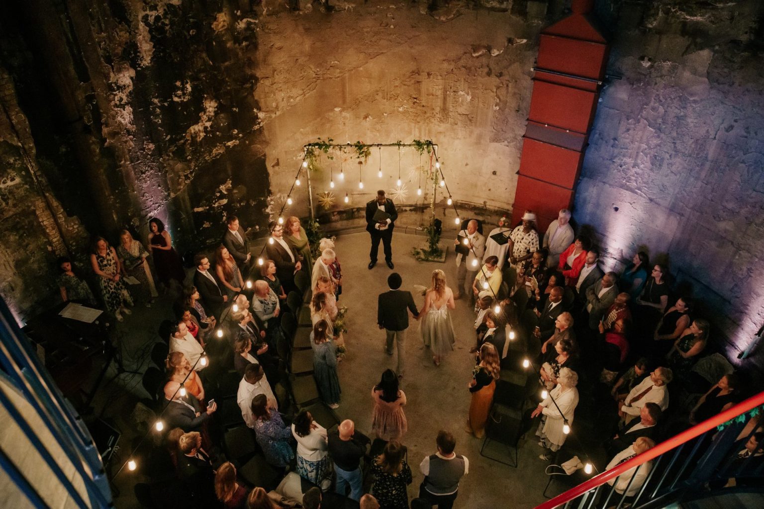 Bride and groom holding hands in front of a registrar for their wedding at Brunel Museum Tunnel Shaft. The background is concrete walls with festoon lights and guests standing in a semi circle