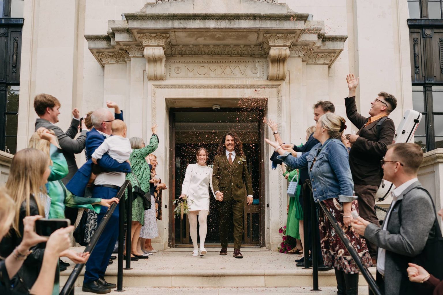 bride in a short white wedding dress and gucci white tights and groom in khaki tweed suit leave islington town hall ceremony and get showered in petal confetti by the guests