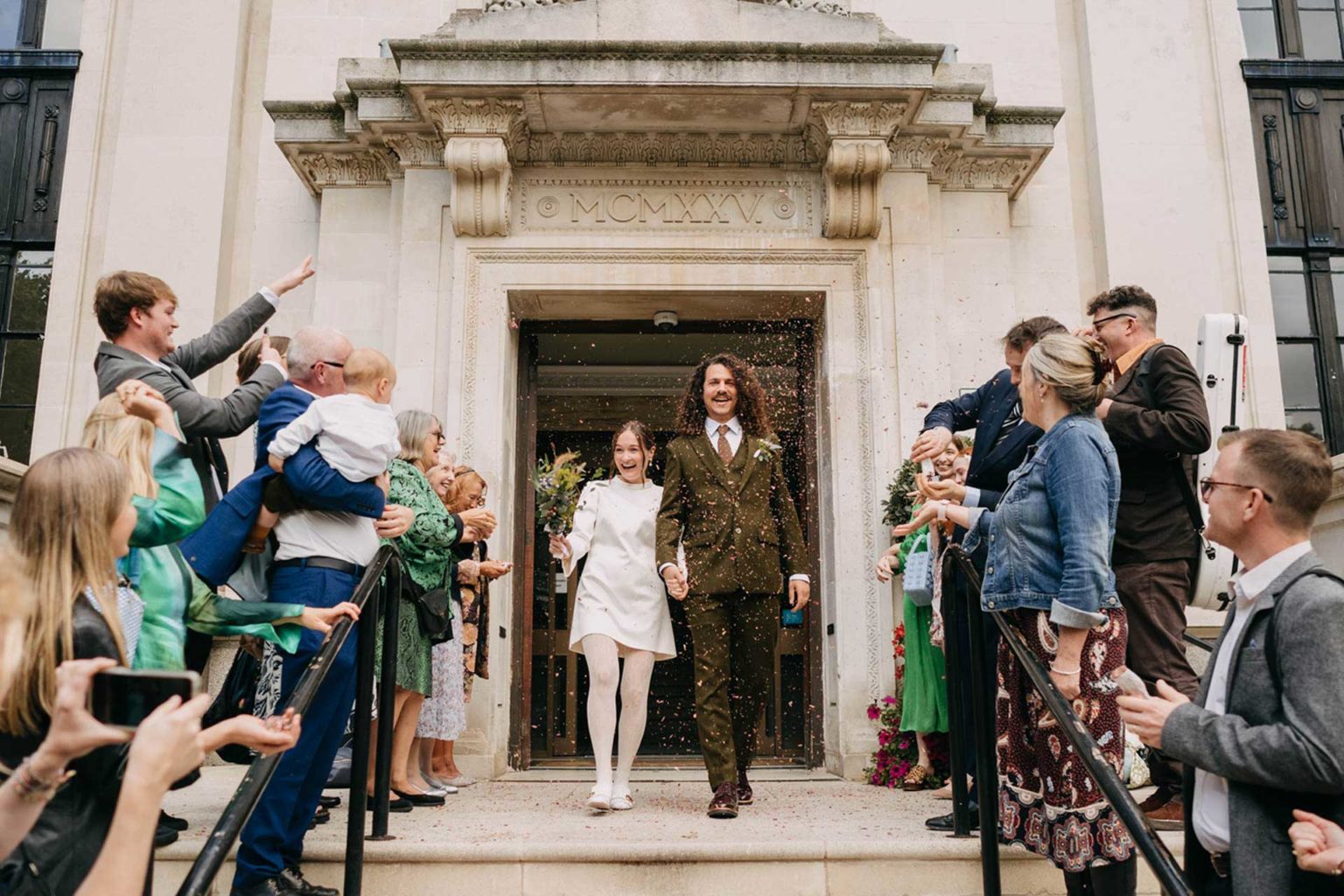 best wedding photography confetti exit at islington town hall stairs with bride in white short dress and groom in olive suit with long curly hair