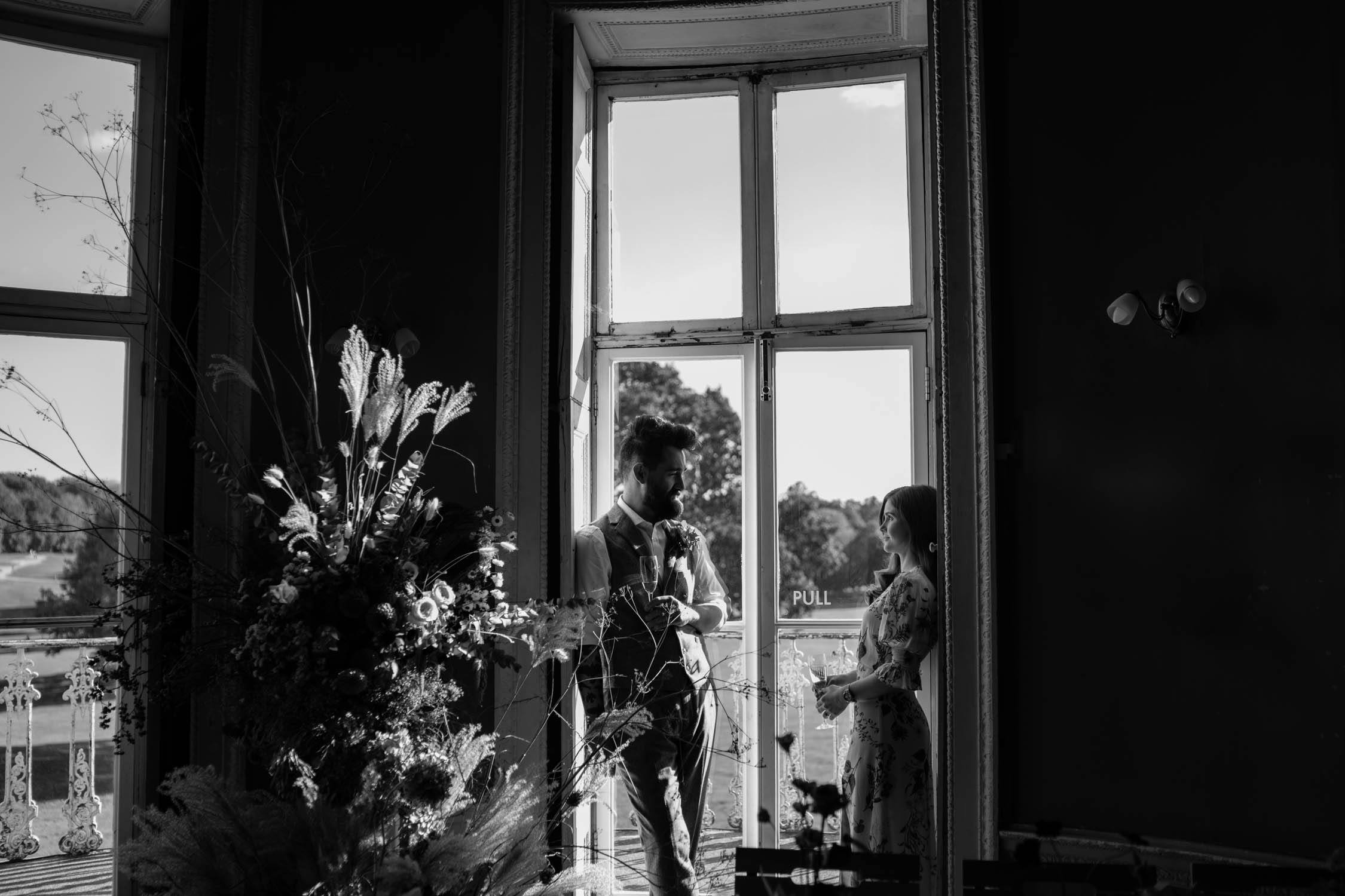 black and white photo of bride and gggroom leanning on a window of beckenham place mansion