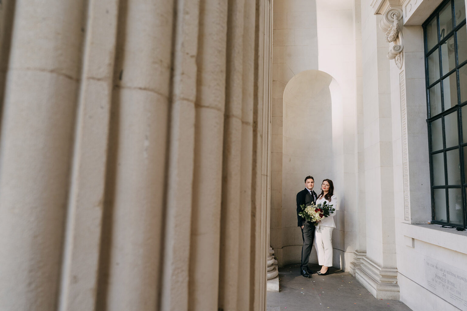 bride and groom posing at the old marylebone town hall colonnade. brde in white luxe suit with large textured bouquet 