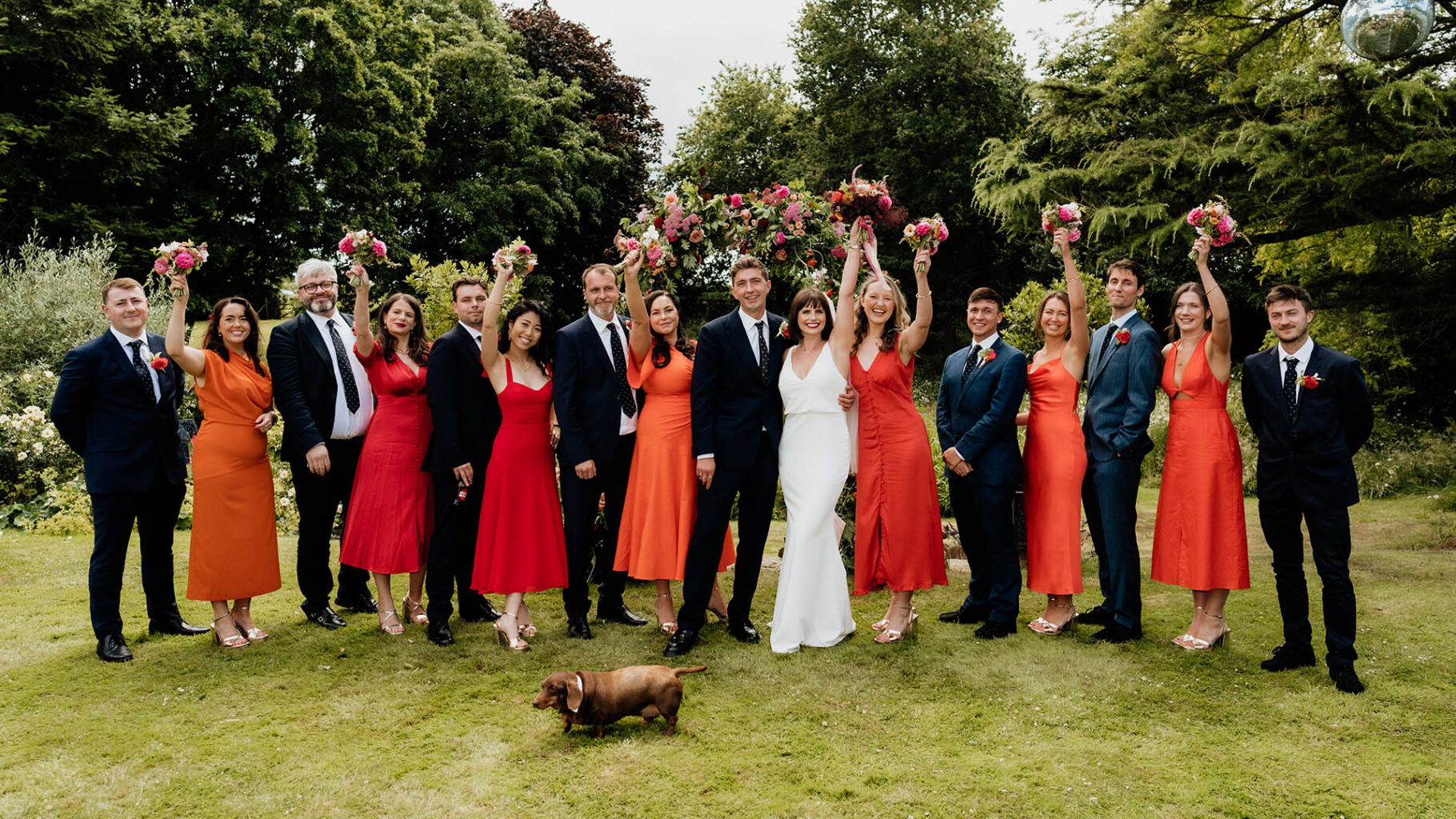 bridesmaids in orange and red dresses waiving bouquets and groomsmen in navy suits