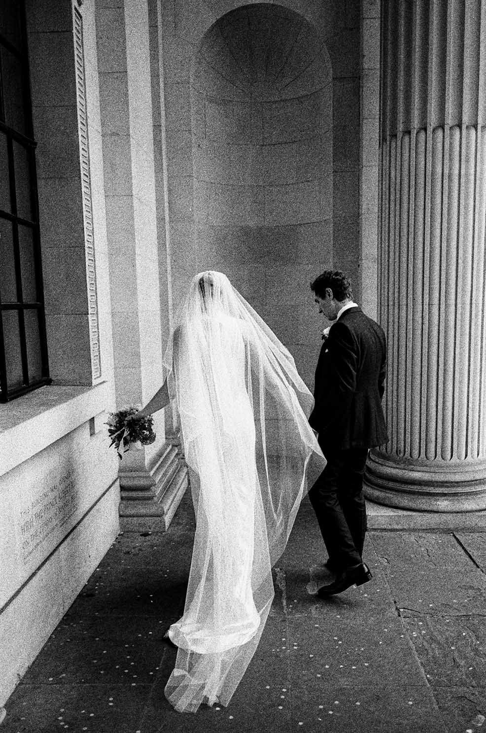 Bride with long veil and groom walking outside the old marylebone town hall captured on black and white film
