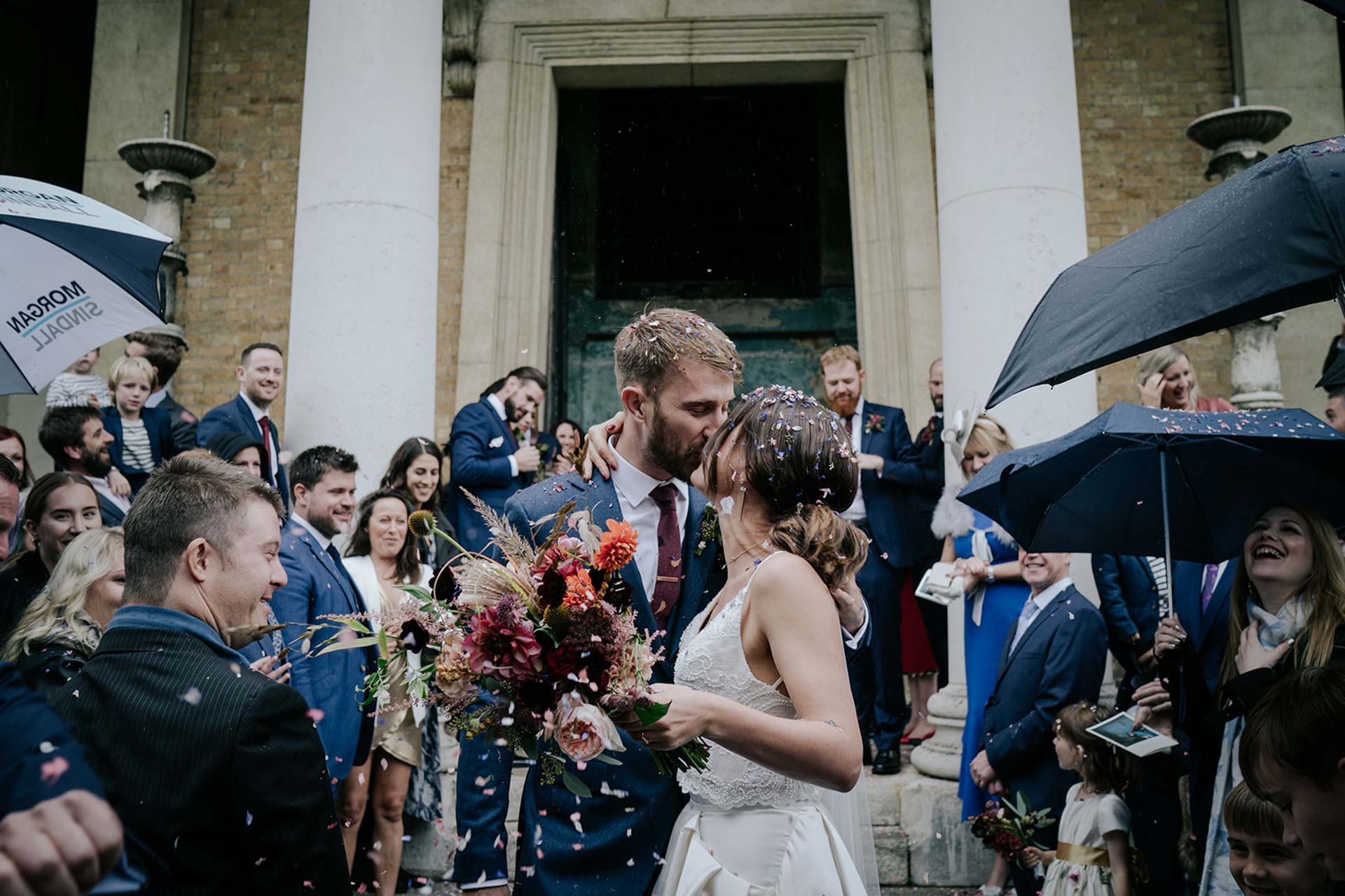 bride and groom kissing at confetti outside asylum chapel in pekcham london surrounded by guests with umbrellas