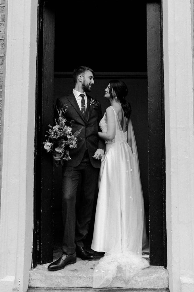 artistic portrait of couple posing in a doorway with bride leaning into groom who is holding her flowers. black and white cinematic photo