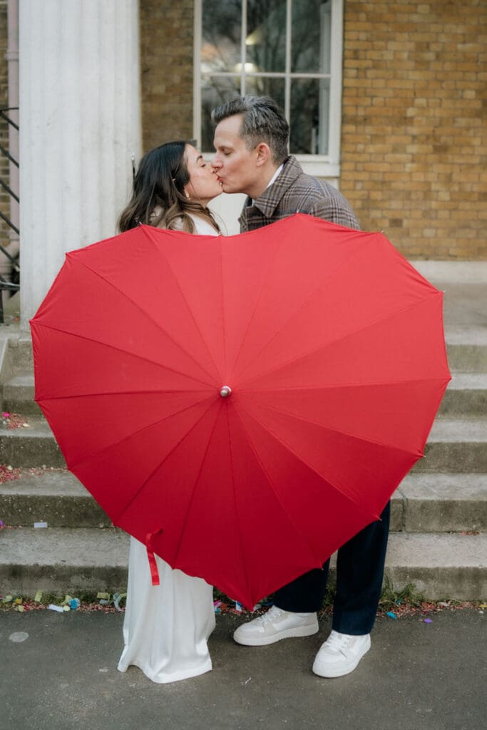 bride and groom kissing behind giant red heart umbrella on a cold rainy wedding day