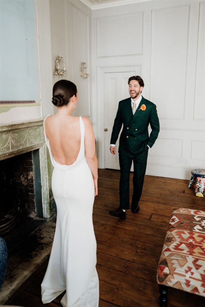 first look with groom in green suit showing happiness upon seeing his bride in white dress with open back -editorial wedding photography