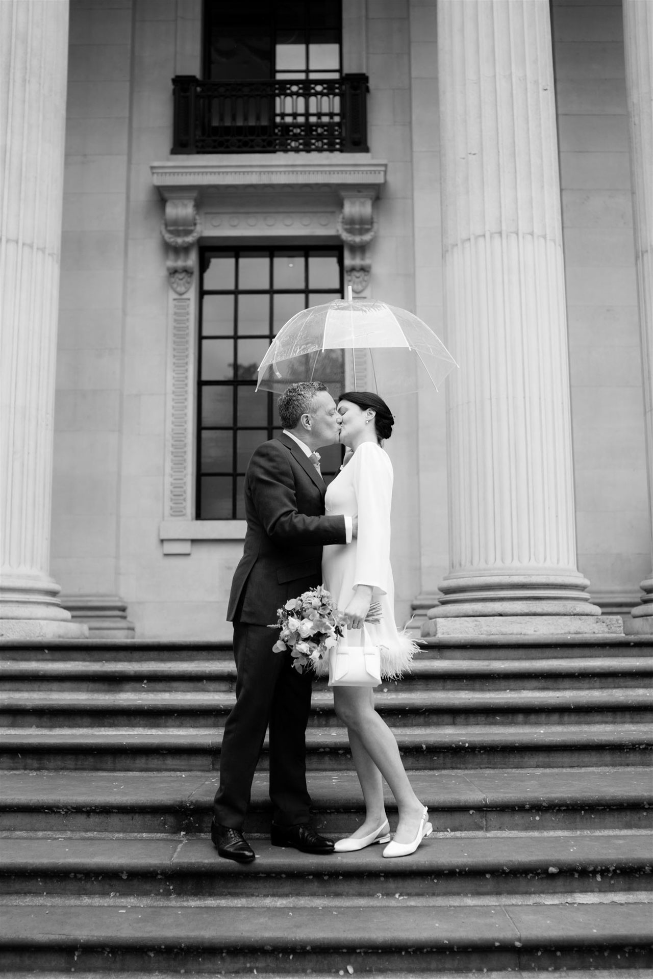 groom kissing bride under clear dome umbrella on stairs of london town hall on a rainy wedding day