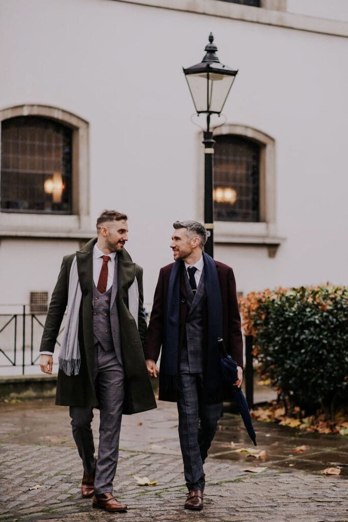 two grooms in warm coats and with scarves, walking holding hands and smiling despite cold wedding day in london
