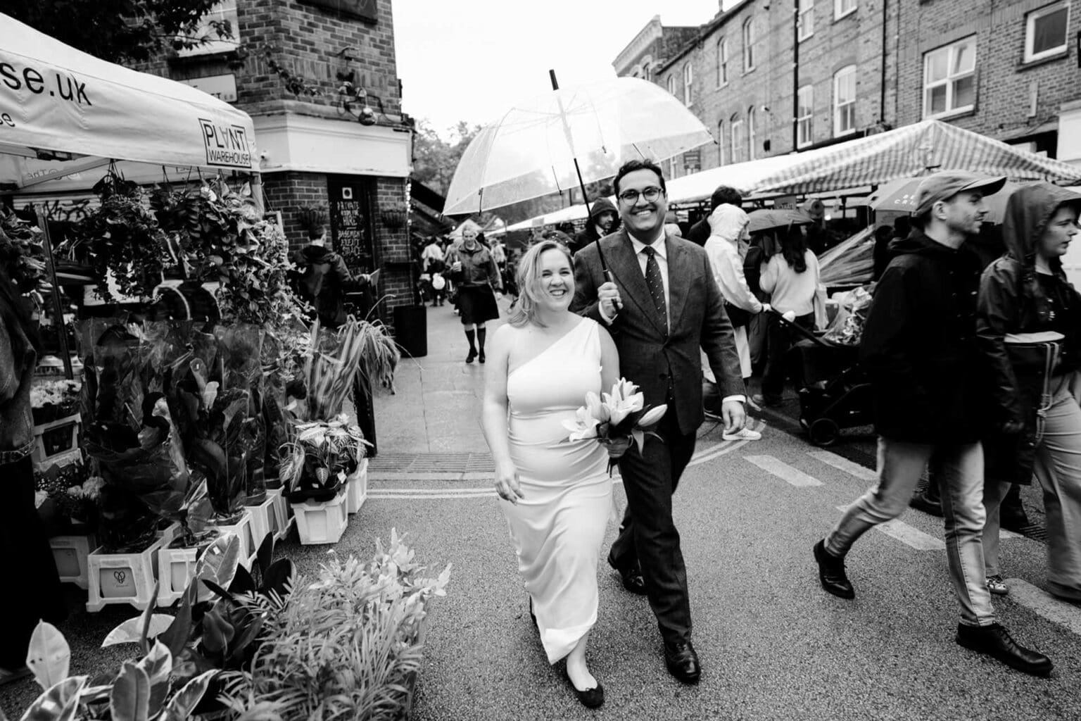bride in flat ballerinas walking with groom holding umbrella over them on a rainy wedding day in hackney london