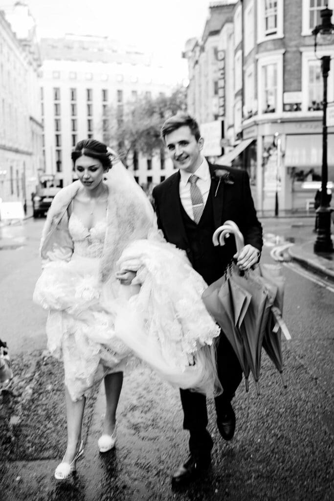 bride holding her dress up with groom carrying umbrellas walking on a rainy wedding day in London