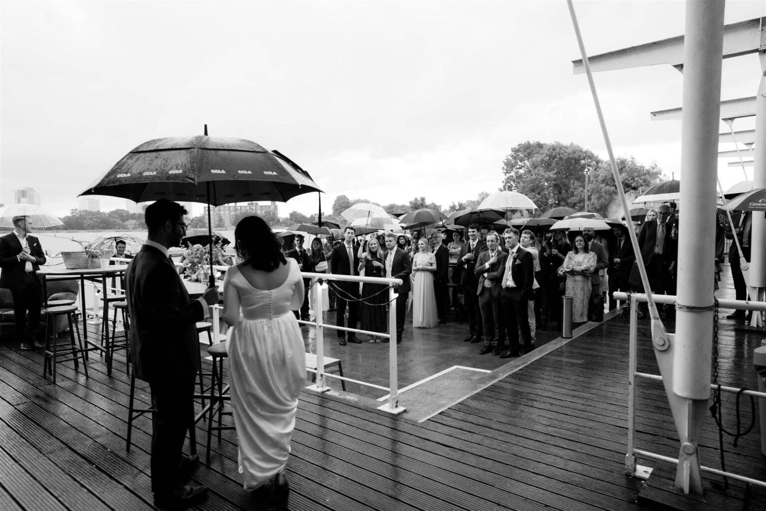 outdoor speeches in rain with bride and groom under a giant umbrella and guests grouped under more umbrellas at london wetlands venue