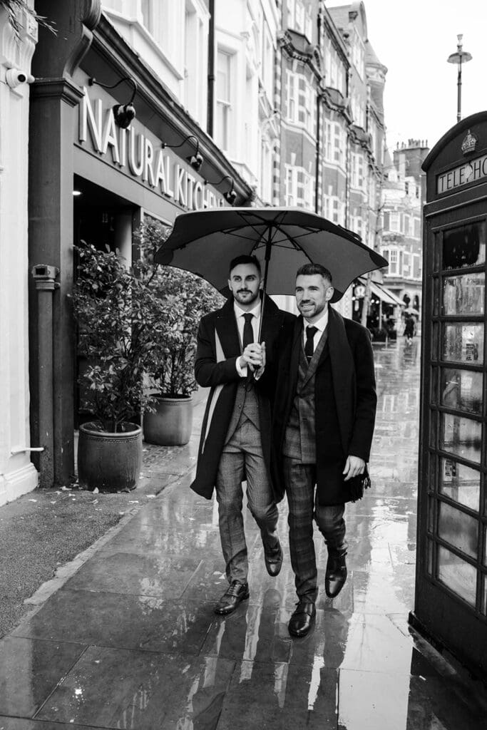 two grooms walking on London streets under an umbrella with wet pavement reflections caused by rain on wedding day