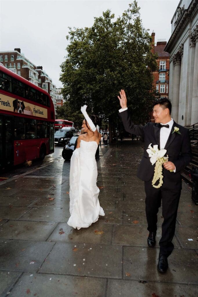 bride and groom waving at guests on a wet wedding day outside the old marylebone town hall with dark skies and wet floor