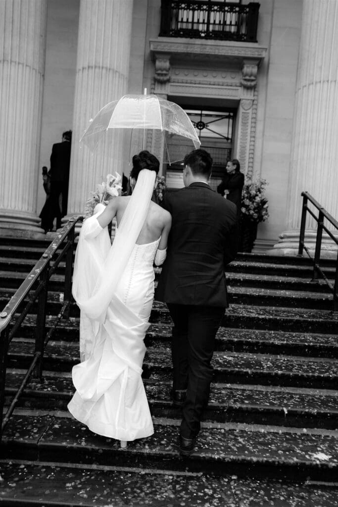 moody cinematic candid photo of bride and room walking up the stairs of the old marylebone town hall under a clear umbrella to shelter from rain