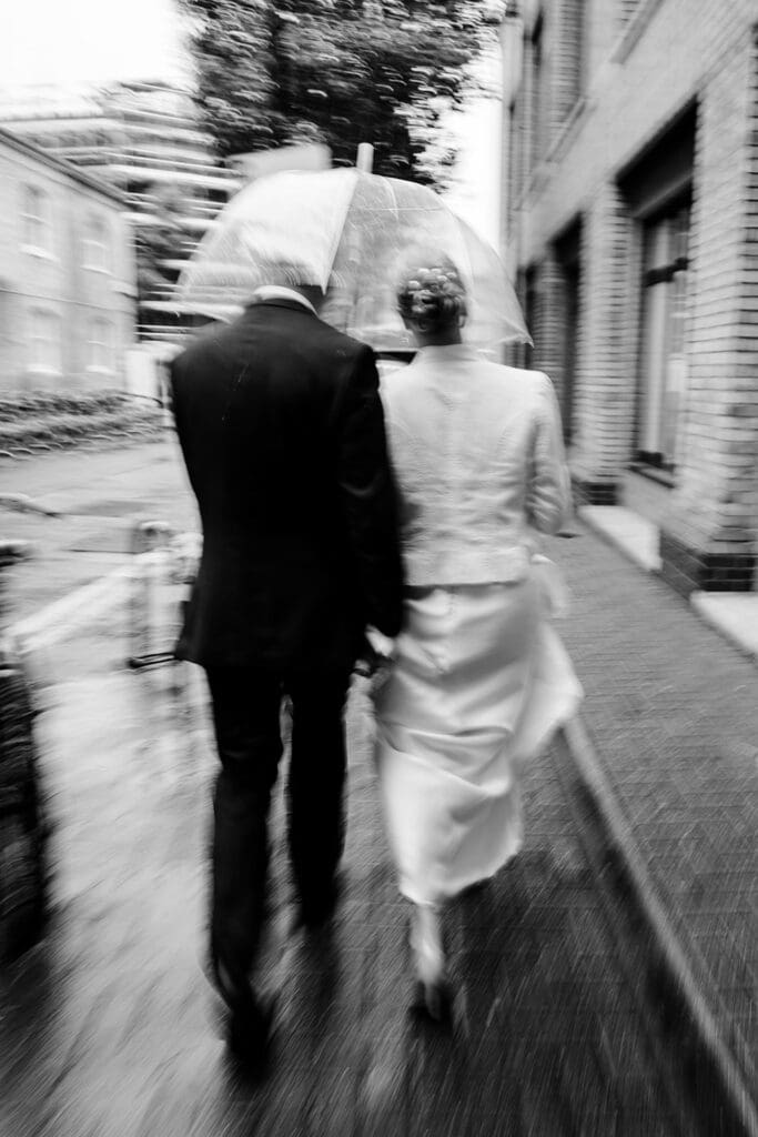 blurry, cinematic photo of bride and groom walking away under a clear umbrella with rain on a wedding day in london