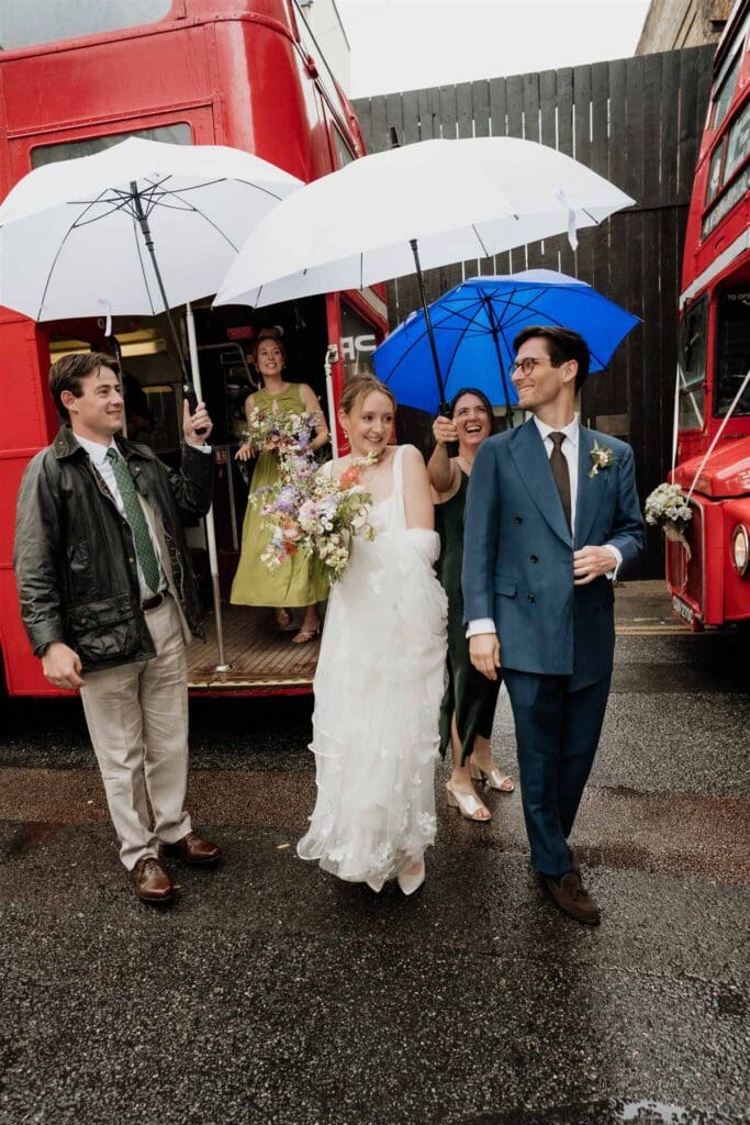 bride and groom with guests leaving a london routemaster and hiding from rain on wedding day with giant umbrellas, smiling