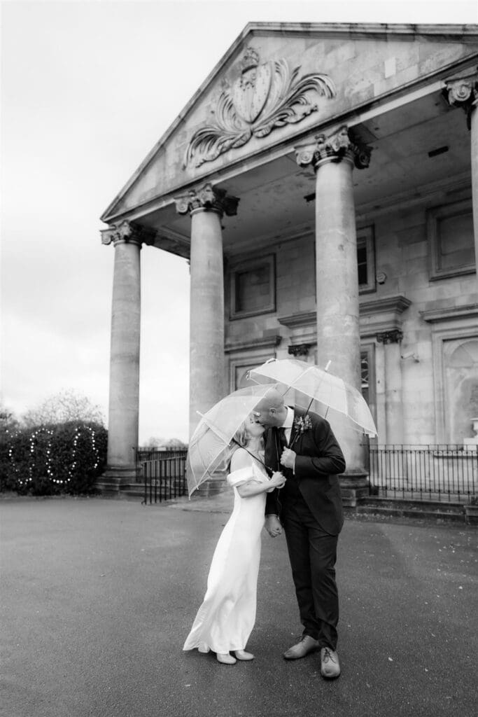 bride and groom kissing under clear umbrellas to protect from rain on wedding day at beckenham place mansion