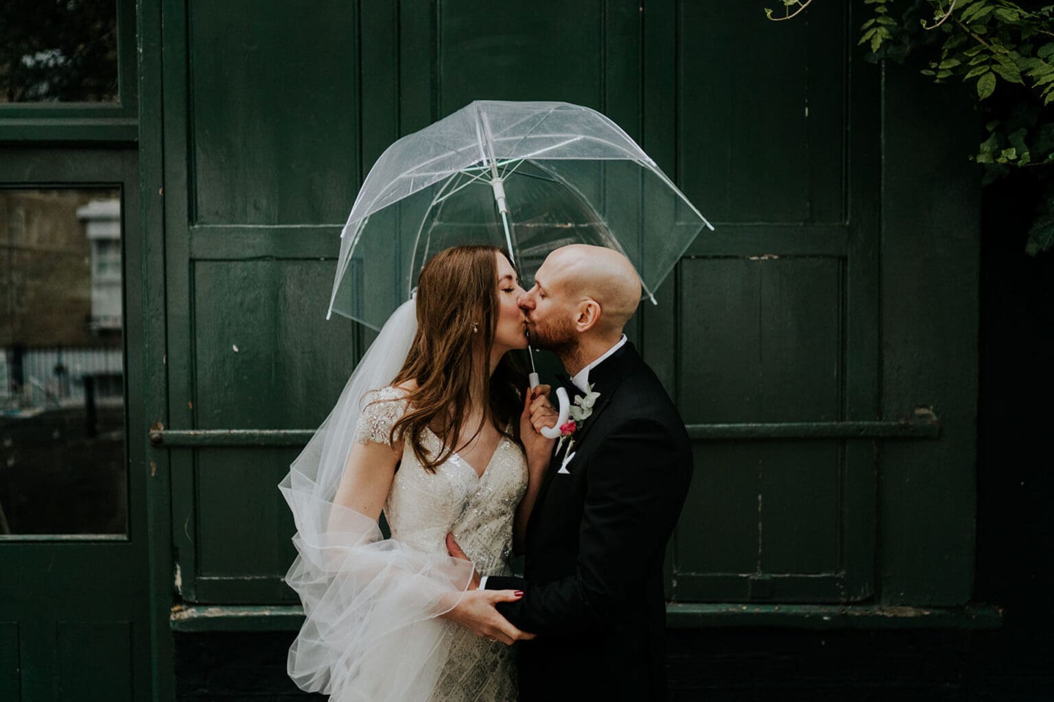 moody cinematic portrait of bride and groom kissing under clear umbrella with green wall behind in islington london