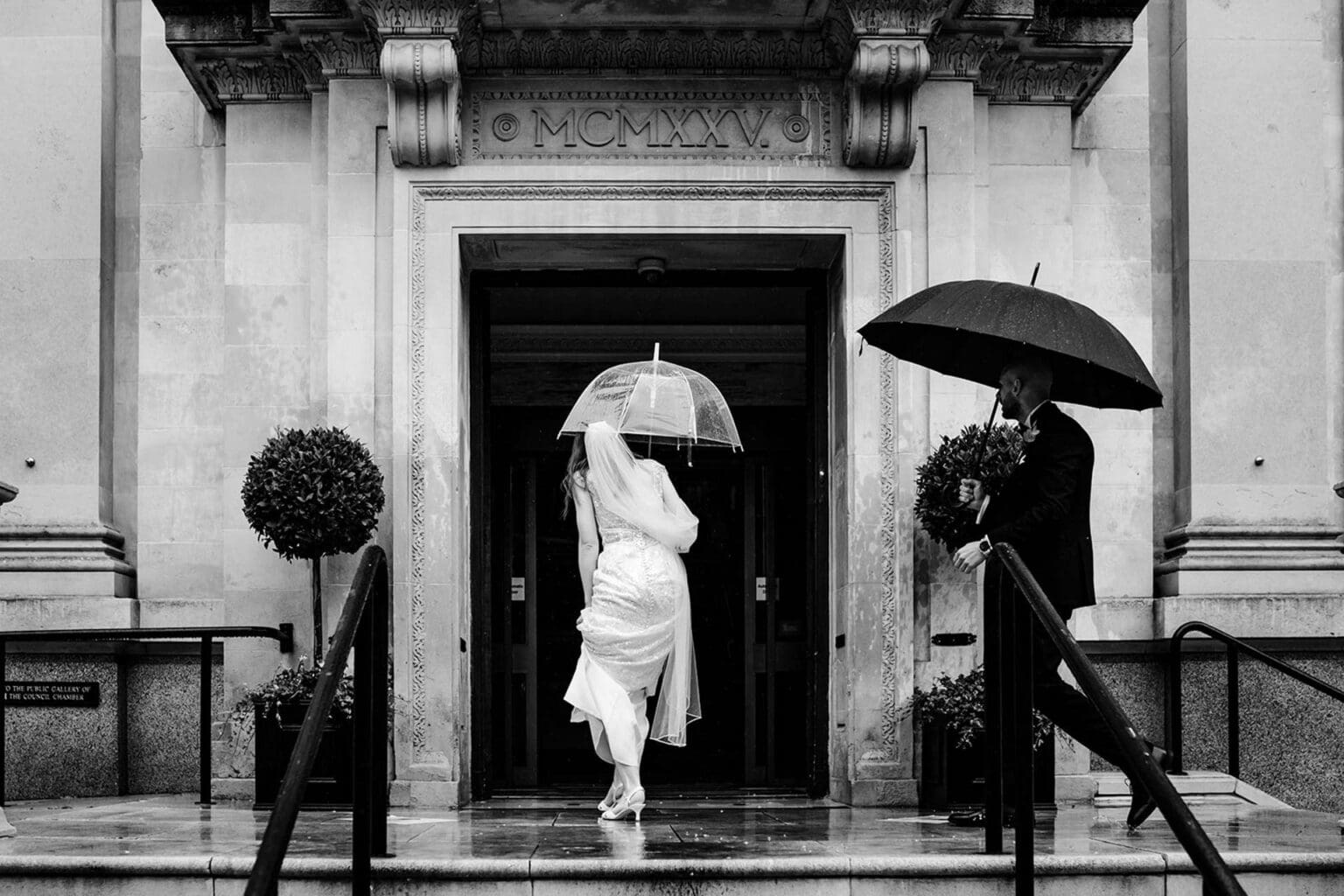 rain on a wedding day with bride and groom holding cleaar and black umbrellas and walking into islington town hall