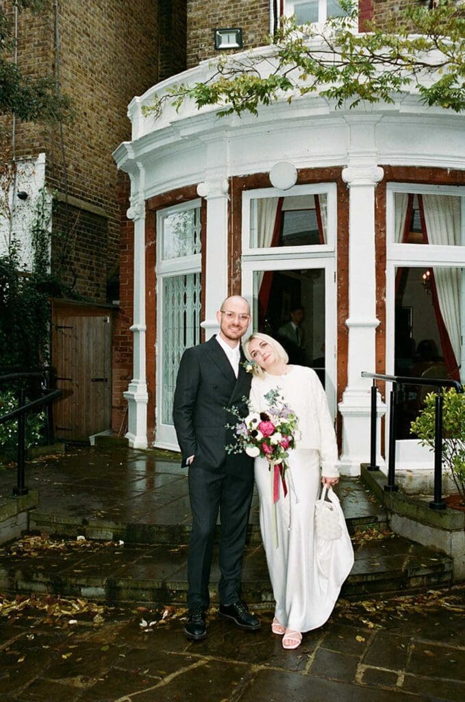 bride leaning on groom and holding her dress on a rainy wedding day at southwark register office in south london, film wedding photo