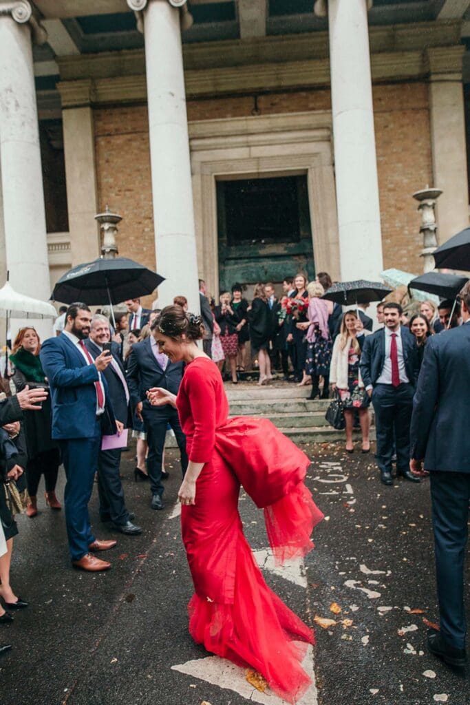 bride in unique designer red dress is laughing after confetti in rain on her wedding day at asykum chapel in london
